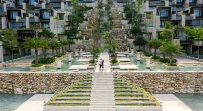 A couple walks through a modern resort with lush gardens and water features, showcasing luxurious architecture.