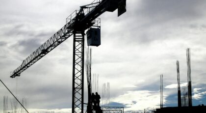 Silhouetted construction crane and workers against a cloudy sky during the day.