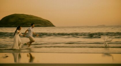 A couple runs joyfully on a beach at sunset with a scenic view of Rio de Janeiro.