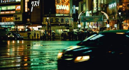 City lights reflecting on wet streets in Shibuya, Tokyo, with blurred traffic and pedestrians carrying umbrellas.