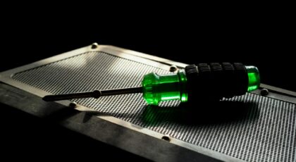 A green screwdriver with a black grip resting on a metallic perforated surface in a studio setting.