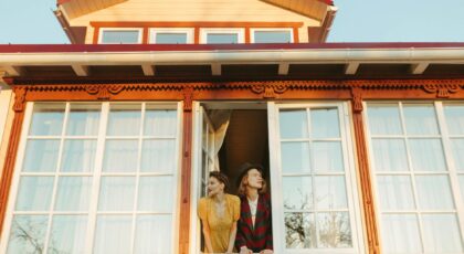 A couple in vintage attire leans out of a picturesque window under a clear sky.