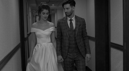 A newlywed couple walks hand in hand through a hotel corridor in black and white.