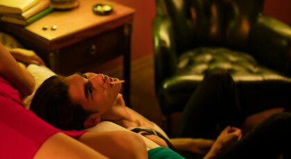 A relaxed couple indoors, reclining on a bed, with a cigarette in dim, warm lighting.