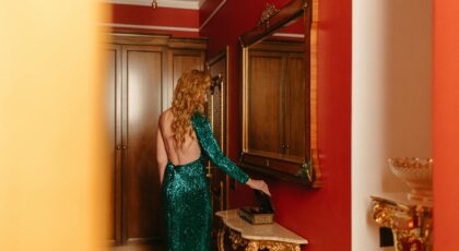 Woman in a sparkling dress admires herself in a mirror inside a luxurious room with rich decor.