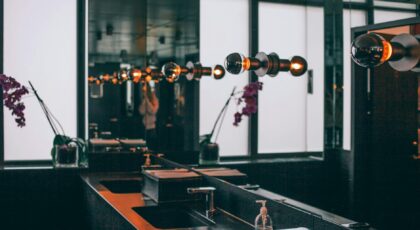 Black sinks with stainless steel faucets placed under wide mirror decorated with illuminated dark lamps in modern toilet room