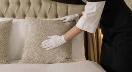 A housekeeper wearing gloves arranges pillows on a luxurious bed in a hotel room.