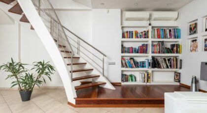 Assorted textbooks on shelves against contemporary staircase above ceramic floor with potted plant and carpet at home
