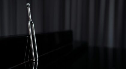 A close-up of a metal tuning fork standing on a reflective piano surface with dark background.