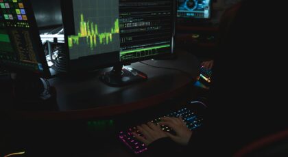 A cybersecurity expert in a dimly lit room is typing on a colorful keyboard with multiple screens displaying data.