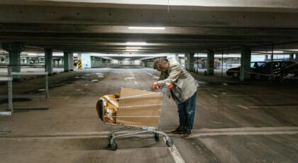 A solitary man leans on a shopping cart in an empty parking garage, evoking a sense of loneliness and urban struggle.