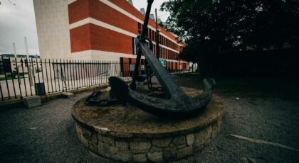 A historic anchor surrounded by gravel and fence in a Dublin park, facing a modern building.