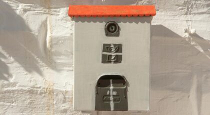 Artistic close-up of a decorative mailbox with a red roof on a textured white wall.