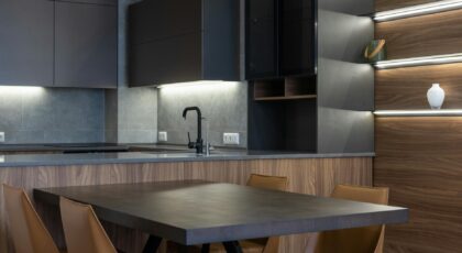 Contemporary black cupboards with sink in stylish kitchen near wooden table and chairs placed near wall with light decoration at home