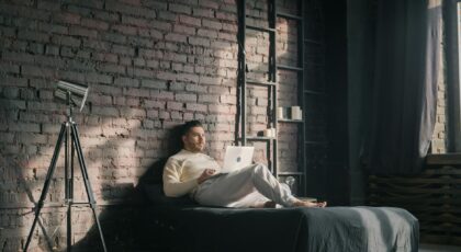 A man lounging on a bed using a laptop in a modern loft setting with natural light.