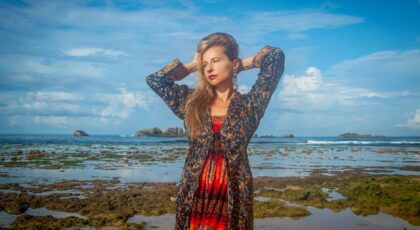 A stylish woman poses on a tropical beach in vibrant attire under a blue sky.