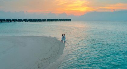 Aerial view of a couple embracing on a sandy beach at sunset with overwater bungalows in the background.