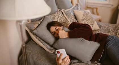 A young woman lying on a bed, holding a phone and a pillow, symbolizing relaxation and connection.