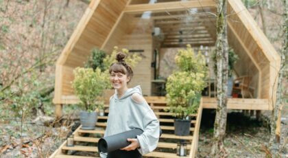 Cheerful woman with yoga mat in front of a wooden cabin surrounded by nature.