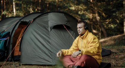 A man enjoys quiet meditation outside a tent in a forest, wearing headphones and a yellow jacket.