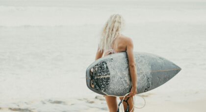 An adult woman carrying a surfboard walks towards the ocean on a sunny day in Bali, Indonesia.