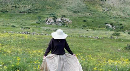 A woman in a sun hat wanders through a lush mountain meadow during the day.