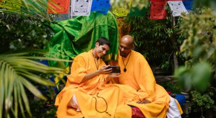 Two monks in bright robes share a moment with smartphone, surrounded by lush greenery and colorful prayer flags.