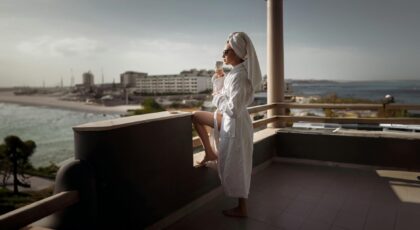 A woman in a bathrobe enjoys a drink on a sunny balcony overlooking the ocean.