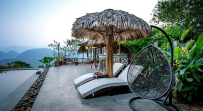 White loungers and thatched parasol with chair placed near swimming pool on terrace of tropical hotel with green exotic plants against mountain ridge on background