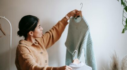 Side view of young pensive female in trendy cardigan looking at knitted sweater and shirt on hangers in cloakroom