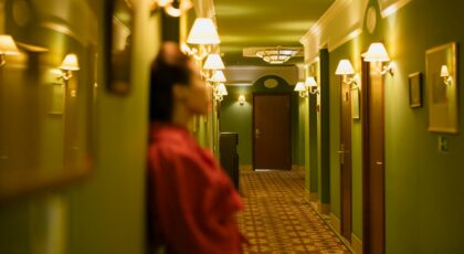 A woman in a red dress leans against the wall in a dimly lit hotel hallway.