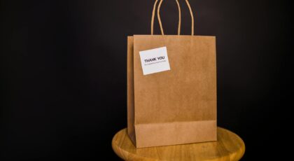 Brown paper bag with thank you note placed on a wooden stool against a black background.