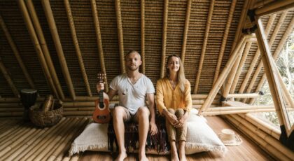 A couple sitting indoors in a bamboo villa, holding a ukulele, enjoying a rustic lifestyle.