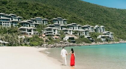 A couple walks hand in hand along a scenic beach next to luxurious villas, enjoying a sunny day.